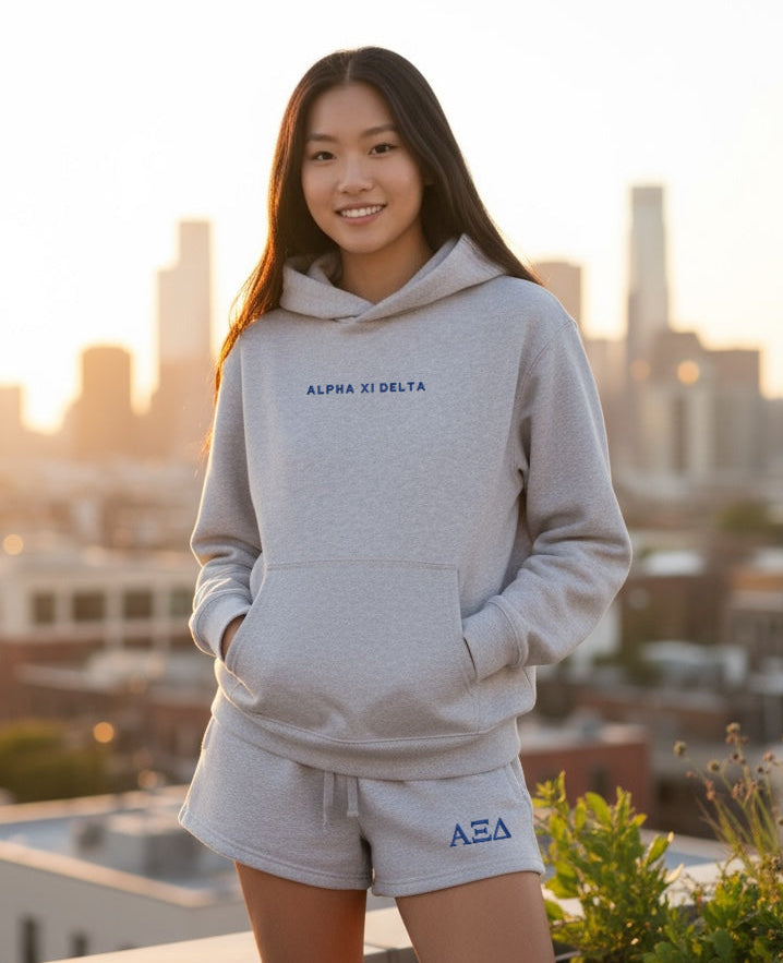 Person wearing a gray hoodie and shorts with 'Alpha Xi Delta' logo, standing on a rooftop with city skyline in the background.
